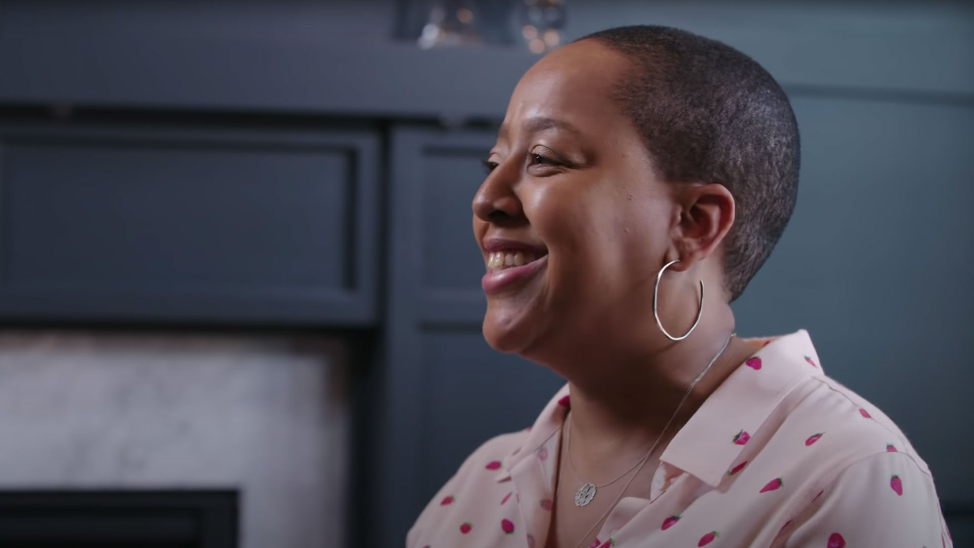 Lesley Pinckney, an Executive Strategy Director and a black woman, sits next to a fireplace in a beautifully designed home. She is wearing a light pink collard dress shirt that has dark pink strawberries.  